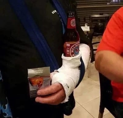 这是一张原始拍摄的静态单图，画面展示了一位手臂打着白色石膏、用蓝色挂带将手臂悬挂在脖子上的人，他巧妙地将一瓶万宝路品牌的啤酒放置在石膏与手臂之间的空隙中，同时另一只手拿着一盒倒置的万宝路香烟。场景看起来是在室内的休闲场所，旁边还能看到另一位穿红色上衣的人的部分身体。这张照片通过这种受伤后依然想尽办法享受烟酒的搞笑操作，营造出一种幽默诙谐的氛围，展现了一种乐观甚至有点硬核的生活态度。