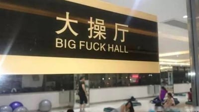 这是一张健身房内的实拍图，画面上方是一块黑黄配色的标识牌，白色字体标注中文“大操厅”，下方的英文翻译却错误地使用了不雅词汇“BIG FUCK HALL”，完全偏离了“健身操大厅”的正确含义，属于典型的翻译失误。标识牌下方的场地中，有几位顾客正在进行拉伸活动，场地内还摆放着健身球等器材，能看出这是健身房内供团体健身课程使用的区域。这张图的笑点在于错误翻译造成的尴尬又搞笑的效果，将原本正常的场所名称变成了不雅表述。