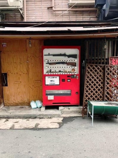 这是一张在日本市井小巷中实地拍摄的照片，画面核心是一台被改造的红色自动售货机，打破了人们对售货机售卖饮料零食的固有认知，原本用于摆放饮品的货道中，赫然陈列着两把长枪类枪械，下方货道还摆放着一些小型物品。售货机正面印有日文标识“野味专门店 KIMONO”，表明其售卖的是和狩猎、野味相关的合规用品。售货机位于一处临街的门口区域，左侧是木质大门，右侧是棕色格栅围栏，地面是有破损痕迹的水泥路面，售货机旁还放着两个白色袋子，右侧有一个绿色小推车，整体场景充满了生活化的市井气息，而售货机内的枪械与日常售货机的反差，制造出强烈的违和感，显得十分新奇。