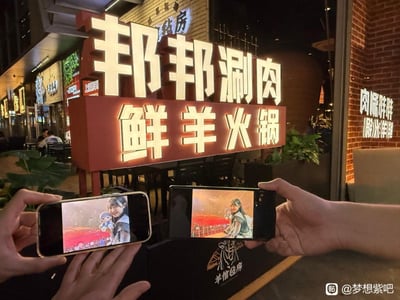 夜晚的街道上，一家名为"邦邦涮肉 鲜羊火锅"的餐厅招牌格外醒目，红色底版配上白色立体发光字，在夜色中十分亮眼。两只手分别拿着智能手机举在招牌前，手机屏幕上显示着动画角色的图片，似乎是在让虚拟角色与现实中的餐厅招牌合影。背景中可以看到餐厅内的灯光和部分就餐环境，整体氛围充满生活气息与二次元文化的有趣结合。