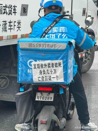 这是一张街头实拍图，画面主体是一位身着饿了么买药配送制服的外卖骑手，正骑着悬挂北京牌照（QT6289）的电动车，处于配送途中。他的蓝色外卖箱上贴有一张手写的硬核警示标语：“发现偷外卖者砍死 身负几十万 亡命狂徒一个”，用夸张戏谑的狠话来防止自己的外卖餐品被盗，背景可见一辆厢式冷藏货车。整张图用搞笑又无奈的方式，吐槽外卖配送中常遇到的餐品被盗问题，风格硬核又接地气。