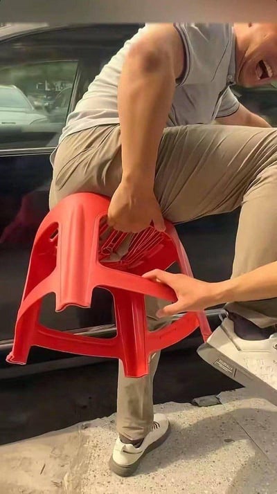 这是一个利用视觉错位打造的整蛊场景：画面中一名穿着浅灰无袖上衣、卡其色长裤的男子，大张着嘴露出痛苦的表情，身体姿态看起来像是被红色塑料凳子牢牢卡住，另一只手还有人在拽动凳子。实际上这是通过特定拍摄角度制造的视觉错觉，凳子并没有真的卡住男子，只是拍摄手法营造出的搞笑假象，整体画面充满趣味，带着整蛊的滑稽感。