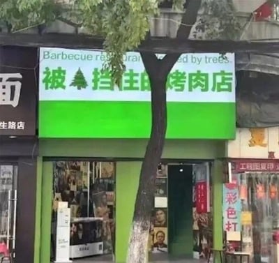 这是一张街头实拍照片，画面主体是一家烤肉店的招牌。招牌分为上下两部分，上方白底黑字写着英文“Barbecue restaurant blocked by trees”，下方绿底白字写着中文“被树挡住的烤肉店”，而一棵大树的树干恰好从招牌正中间穿过，完美呼应了店名的描述，将现实中的遮挡情况变成了店名的创意点，趣味十足。招牌两侧还有其他店铺的标识，左侧是带“面”“生路店”字样的店铺，右侧有“工程图复印”“彩色打”字样的店铺。