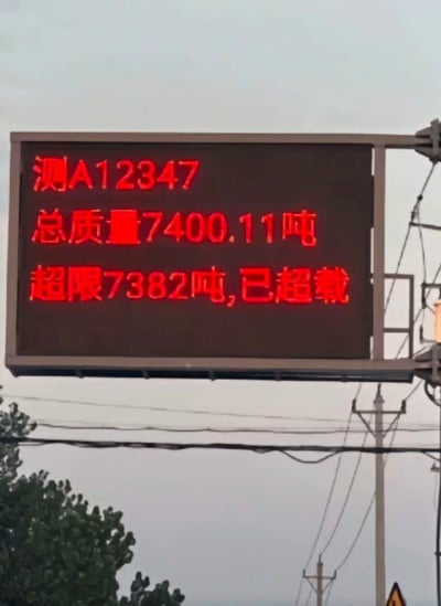 图片中是一块户外电子显示屏，屏幕上用红色字体显示着车辆信息：车牌号为“测A12347”，总质量7400.11吨，超限7382吨，并标注“已超载”。背景可见电力线和树木，推测拍摄于道路旁。超限数值高达7382吨，远超正常货车载重范围，明显存在系统故障或数据错误，形成荒诞搞笑的效果。