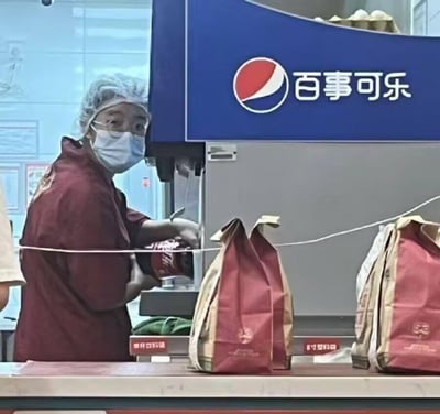 图片中一位身着红色工作服、佩戴发网和口罩的百事可乐员工，正站在带有明显百事可乐标志的蓝色背景板前。令人意外的是，她手中竟拿着一罐红色的可口可乐，与身后的品牌标识形成鲜明对比。柜台上还摆放着几个红色的纸袋，整体场景充满了品牌错位的幽默感。