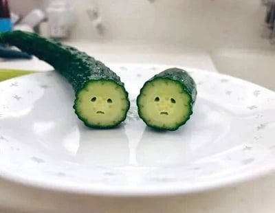 这是一张P图改编的静态单图，画面主体是被切成两段的黄瓜，放置在带有浅淡花纹的白色盘子上。创作者在两段黄瓜的切面上用简单的线条画出了下垂的眉眼和小嘴巴，赋予了黄瓜委屈、难过的拟人化表情，仿佛它们因被分开而感到伤心，整体风格轻松搞笑，通过拟人化的手法为普通的食材增添了趣味性，给人一种治愈又滑稽的观感。