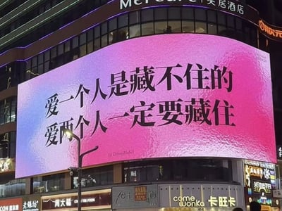 夜间城市商业区域的大型LED广告牌，背景为粉紫渐变色调，中央醒目地显示着黑色文字：“爱一个人是藏不住的 爱两个人一定要藏住”。广告牌周围可见“美居酒店”（Mercure Hotel）、“周大福”珠宝店、“卡旺卡”（Come Wonka）饮品店等商业招牌，整体呈现出繁华的都市夜景氛围。文字内容通过对比“爱一个人”与“爱两个人”的不同处理方式，形成一种幽默且略带讽刺的情感调侃效果。