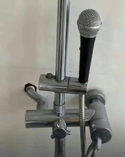 图片展示了一个创意DIY改造场景：银色金属材质的花洒固定支架上，被巧妙地加装了一个黑色手持麦克风。花洒本体的管道、调节旋钮和淋浴软管清晰可见，而麦克风则通过支架夹固定在花洒立杆上，形成一种“浴室K歌设备”的幽默视觉效果。整体构图简洁，背景为纯白色墙面，突出改造后的装置主体，给人一种脑洞大开的创意感。