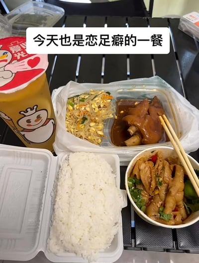 图片展示了一份丰富的外卖餐食，包括白色餐盒中的米饭、透明盒内的豆芽小菜与红烧猪蹄、以及一碗凉拌鸡爪，旁边配有一杯蜜雪冰城的橙色饮料。上方文字“今天也是恋足癖的一餐”使用了谐音梗，将“恋足癖”幽默化，实际指餐食中包含猪蹄和鸡爪（均为动物足部），营造出轻松搞笑的氛围。