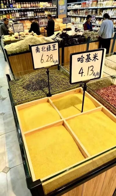 超市粮食区货架实拍图，多个透明玻璃柜内整齐摆放着绿豆、黄色谷物等粮食。前景中两个黑色边框白色底板的价格牌清晰可见：左侧标注'南北绿豆 6.28元/斤'，右侧则标有网络流行词'哈基米 4.13元/斤'，将粮食名称替换为萌宠代称制造反差笑点。背景中几位顾客正在挑选商品，货架上层陈列着各类瓶装调料和袋装食品，整体场景真实生活化，因'哈基米'的趣味标注形成独特的网络文化与现实场景结合的幽默效果。