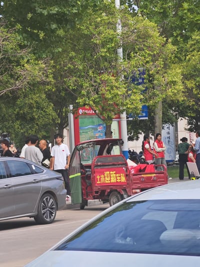 校园街道场景中，一辆红色三轮车停在路边，车身上醒目地写着“土木迎新车队”字样。周围有多位行人，包括穿着白色T恤的男生和几位身着红色马甲的志愿者，背景是茂密的绿树和一块校园导览牌。道路上还有几辆轿车驶过，整体氛围热闹，像是大学新生报到期间的迎新场景。