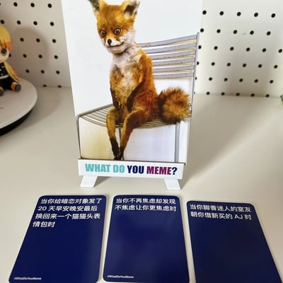 图片展示了What Do You Meme?桌游套装的桌面场景：中央立着一张印有狐狸直立坐在椅子上的卡牌，卡牌下方标注"WHAT DO YOU MEME?"；卡牌前方摆放三张蓝色 caption 卡，分别写着“当你给暗恋对象发了20天早安晚安最后换回来一个猫猫头表情包时”“当你不再焦虑却发现不焦虑让你更焦虑时”“当你脚香迷人的室友朝你借新买的 AJ 时”。背景为白色钉板，左侧有一个小玩偶。