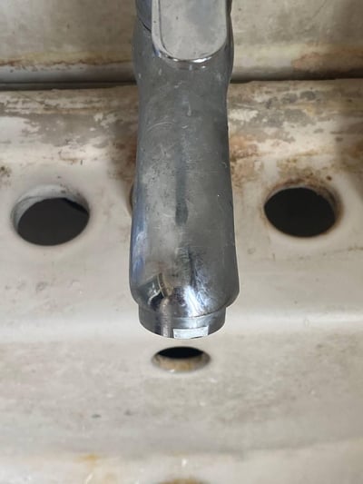这是一张近距离拍摄的洗手台局部特写，画面中心是一支表面布满划痕、磨损严重的镀铬水龙头，水龙头下方的陶瓷洗手台带有三个预留孔洞，台面上附着泛黄的水垢与污渍，整体呈现出长期使用、缺乏清洁维护的老旧状态。