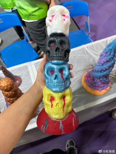 一只手正拿着一个由五个不同颜色骷髅头堆叠而成的手工艺品。骷髅头从上到下分别是白色、黑色、蓝色、黄色和红色，每个骷髅头上都有红色的类似血迹的装饰。背景中可以看到蓝色的椅子和一个穿着绿色上衣的人。这似乎是在某个手工艺品展示或创意市集的场景。