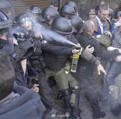 这张图片记录了混乱的冲突现场：一群身着全套防暴装备的警察正和民众发生推搡拉扯，其中一名防暴警察手持催泪喷雾罐，却出现了严重失误——将白色的喷雾直接喷向了自己身旁的队友，而非对面的人群。现场氛围紧张又带着尴尬的荒诞感，周围的警察和民众都处于混乱的拉扯状态。