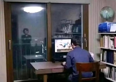 室内场景中，一名男子背对着镜头坐在书桌前，正专注地操作电脑，屏幕上显示着画面；玻璃推拉门的另一侧，一名女子的身影清晰可见，正静静地注视着男子的背影。整个画面营造出一种被窥探、被监视的诡异压迫感，没有双关或谐音梗内容。