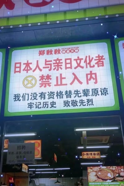 这是一张夜晚街边火锅店的实拍图，店铺名为「郑秋秋创业火锅」，招牌上用醒目的红色大字标注「日本人与亲日文化者 禁止入内」，搭配禁止符号，下方配有文字「我们没有资格替先辈原谅 牢记历史 致敬先烈」，鲜明表达了店铺的立场。店铺下方还能看到招聘启事和「小芳理发店」的标识，整体场景是普通的街边商铺环境。