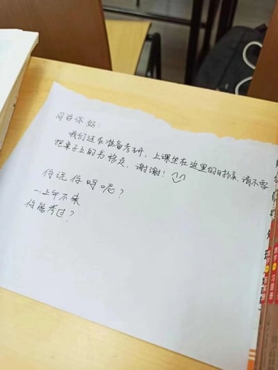这张图片拍摄的是大学校园教室里的一张手写纸条，纸条上包含两段内容：上方是准备考研的同学写下的礼貌请求，他们希望上课的同学不要移走放在桌子上的书，还附带了一个比心的表情，态度友好；下方是其他同学留下的阴阳怪气的回复，质疑备考同学一上午不来自习，能不能通过考研，整体展现了校园里考研占座引发的小摩擦，是真实的校园生活场景的体现。