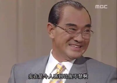 图片为一名中年男性的电视节目截图，他身着灰色西装、白色衬衫，系着黄紫条纹领带，头发梳得整齐，戴着眼镜，正微笑着转头侧脸。背景是纯色米色墙面，底部有中文字幕“实在是令人感到忍俊不禁啊”，右上角可见韩国MBC电视台标志，底部有水印“@电工厨”。