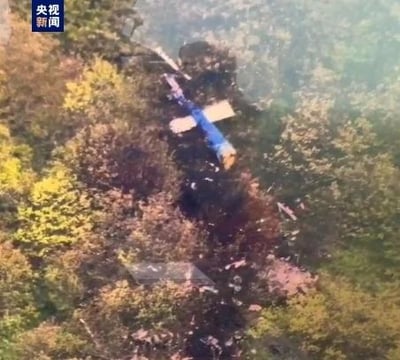这是央视新闻的报道截图，画面呈现了一处山林中的直升机坠毁事故现场：一架蓝黄相间的直升机残骸落在茂密的树林间，机身损毁严重，周边植被有明显的烧灼、破坏痕迹，直观展现了事故的惨烈程度，传递出沉重的事故氛围。