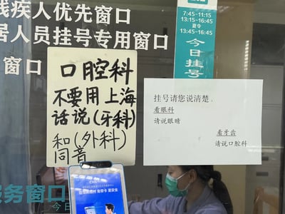 这是一张实拍的上海社区卫生服务中心挂号窗口的照片，窗口玻璃上贴有多张标识和提示：原有标识显示该窗口为残疾人优先、挂号专用窗口，同时标注了普通时段和夏令时的挂号营业时间。另外张贴的两张提示针对挂号易混淆的问题作出说明：左侧手写提示表示上海话里“口腔科”和“牙科”“外科”发音同音，要求挂号时不要用上海话说相关表述避免挂错号；右侧打印提示明确要求挂号时表述清晰，看眼科要说“眼睛”，看牙齿要说“口腔科”。窗口内有一名佩戴口罩的女性工作人员在岗，窗口前还摆放了宣传医保刷脸支付服务的电子设备。