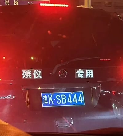 这是一张夜间拍摄的黑色奔驰汽车尾部实拍图，奔驰车标左右两侧分别贴有“殡仪”“专用”的白色标识贴纸，车辆悬挂的天津机动车牌照为“津K·SB444”，牌照中的字母组合与三个连续数字4（谐音同“死”）和“殡仪专用”的标识形成巧合呼应，充满趣味性，画面背景能看到车辆亮起的红色尾灯以及上方路牌的部分文字。