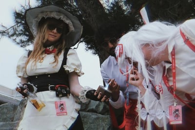 这是一张在东方Project主题展会TouhouFest现场拍摄的趣味cosplay照片：左侧女生cos东方Project里的雾雨魔理沙，戴着墨镜，手持带小烟的玩偶和酒瓶，造型一改原作里的魔法使形象，显得十分“社会”；中间的男生戴着墨镜，正在用打火机点烟；右侧白发coser扮演东方Project的藤原妹红，正低头凑过来点烟，白发和红白服饰还原角色，但抽烟的动作和原作里的不死人形象形成强烈反差。三人在户外松树下，挂着展会的Attendee挂绳，整体氛围搞怪随性，用打破原作设定的方式制造出反差笑点。