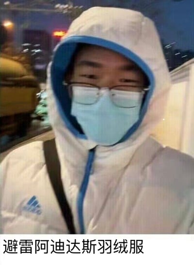 图片中是一位戴着眼镜和浅蓝色口罩的人，穿着白色连帽阿迪达斯羽绒服，内有蓝色内衬。背景为城市夜晚的街道，有模糊的车辆灯光。图片底部有文字“避雷阿迪达斯羽绒服”，疑似对该款羽绒服进行负面评价或吐槽。