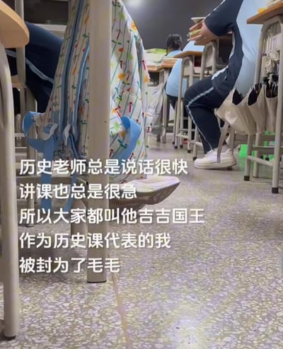图片展示了一个教室场景，从低角度拍摄学生们坐在课桌前的下半身，能看到穿着校服的学生、书包和教室地面。图片上叠加文字讲述了一个校园趣事：历史老师因为说话太快、讲课很急，被学生们戏称为"吉吉国王"，而作为历史课代表的叙述者则被封为"毛毛"。这是一种学生间的幽默调侃，利用了名字的谐音和联想制造笑点。