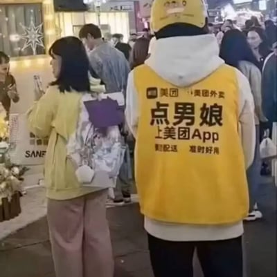 图片中显示一名身穿黄色美团外卖背心的男子站在拥挤的人群中，背心上的文字被修改为"点男娘 上美团App"，是对美团外卖制服的幽默改编。周围环境看起来像是在商场或热闹的公共场所，有许多行人。