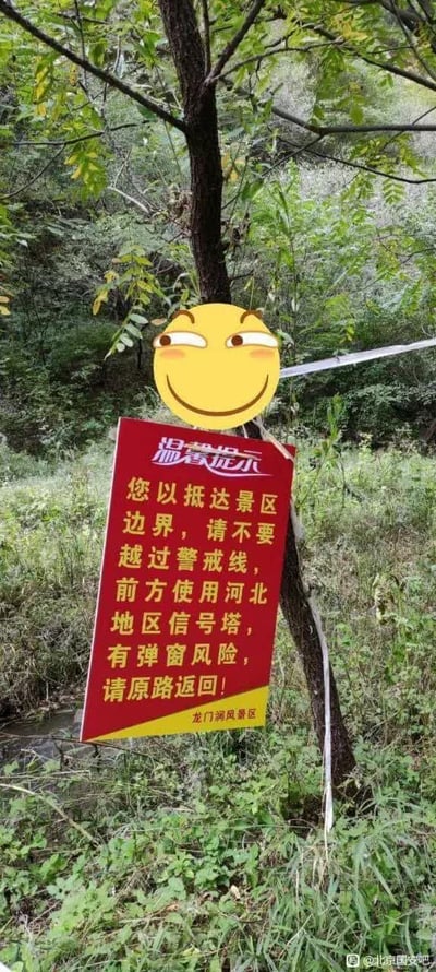 图片展示了龙门涧风景区边界处的一块红色警示牌，背景为茂密的森林植被。警示牌上用黄色文字写着：“您以抵达景区边界，请不要越过警戒线，前方使用河北地区信号塔，有弹窗风险，请原路返回！”，落款为“龙门涧风景区”。牌面上方被添加了一个黄色的滑稽表情emoji，表情眯眼带红晕，显得俏皮幽默。该图片通过将正式的景区提示与网络流行表情结合，形成了一种轻松诙谐的警示效果。