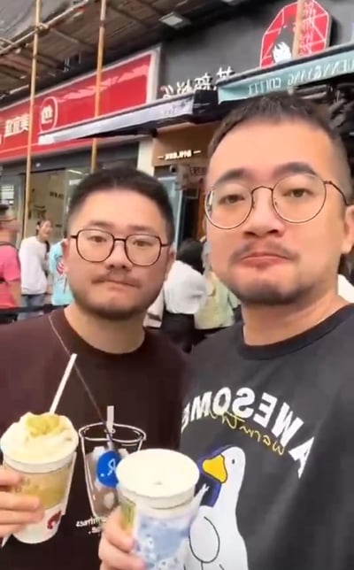 两位戴着眼镜、留着胡须的男士在热闹的街头商铺前自拍，两人各自手持不同的饮品，背景是营业中的店铺和过往路人，整体氛围轻松休闲，是日常出游相聚的生活记录画面。