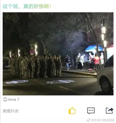这是一张夜晚户外拍摄的实拍照片，画面左侧是一群身着迷彩服的学生，他们整齐地在道路上列队，目光齐刷刷投向路边的烧烤摊；画面右侧的烧烤摊处，摊主正在忙碌制作食物，冒出阵阵烟雾，还有顾客在等待取餐。图片顶部配文“这个班，真的好惨啊！”，通过军训学生只能列队围观他人享用烧烤的反差场景，调侃军训期间的无奈与有趣日常，充满了搞笑的氛围。