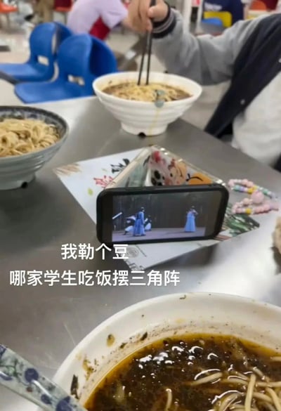 这是一张拍摄于校园食堂的实拍照片，画面展示了学生用餐时的趣味创意：餐桌上摆放着三碗面，其中一碗已食用至只剩汤底，另外两碗还有剩余面条，一名学生正用筷子夹取碗中的面条。为了在吃饭时能观看视频，学生利用两部手机和一张纸搭建出三角形状的简易支架，用来固定播放视频的手机，手机屏幕中正在播放古装类影视内容，旁边还放着一串手机挂链。图片上的配文“我勒个豆 哪家学生吃饭摆三角阵”以调侃的语气，凸显出这个自制支架的脑洞与趣味，充满了校园生活的轻松感。