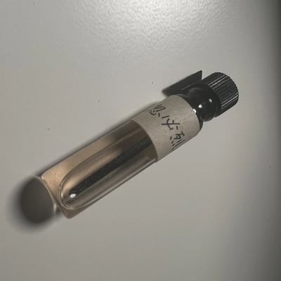 一张展示小瓶子的照片，瓶子放置在白色平面上。瓶子为透明材质，带有黑色瓶盖，瓶身贴有白色标签，上面有手写的中文字符。瓶内似乎装有深色液体或物质。光线从上方照射，在瓶身形成轻微阴影，整体画面简洁干净。