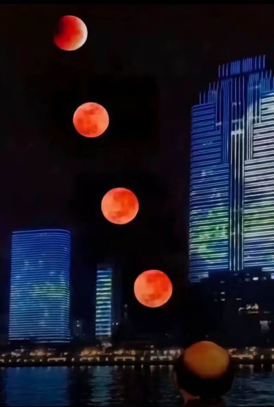 这张图片展示了一个超现实的城市夜景，画面中有四枚红色的月亮悬挂在夜空中，呈现出不同的月相状态。前景中可以看到一个人的后脑勺剪影，正注视着这一奇特的天文景象。背景是现代化的城市建筑群，高楼大厦被蓝色和绿色的灯光照亮，建筑物的轮廓在黑暗中清晰可见。水面倒映着城市的灯光，增添了画面的层次感。整个场景营造出一种神秘而梦幻的氛围。