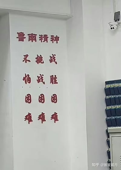 这是一张室内实拍图，墙面张贴着以“鲁南精神”为主题的特殊排列标语：将“不怕困难，挑战困难，战胜困难”三句话拆分后按列排布，横向阅读是“不挑战 怕战胜 困困困 难难难”，竖向阅读则是完整的三句励志口号，形成了有趣的视觉巧思。画面右上角可见监控摄像头，右下角有文件收纳架，同时带有“知乎 @披星戴月”的水印。