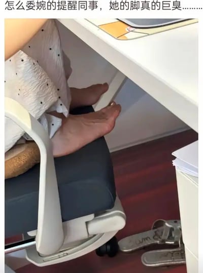 这张图展现了办公室场景，女同事把脚翘在椅子上，鞋随意放在地板上。上方配文直接表达了发帖人苦恼于同事脚气味巨大，想要委婉提醒她，又怕尴尬，十分真实且搞笑。图中也流露出社交困境和职场日常的无奈。配文“巨臭”已经成为网络梗，常用于夸张吐槽。