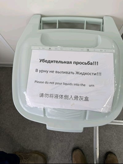 这是一张现场实拍的公共标识错误梗图，画面主体为一个淡绿色的带盖塑料垃圾桶，桶盖表面用透明胶带粘贴了一张三语提示标识：最上方为俄语，内容为“恳请！请勿向桶内倾倒液体！”，中间为英文，内容为“Please do not pour liquids into the urn”，最下方的中文翻译出现严重错误，将英文中表示“垃圾桶、瓮”的“urn”误翻译为“骨灰盒”，最终中文提示变为“请勿将液体倒入骨灰盒”，画面底部还露出了拍摄视角下的几双鞋子，离谱的翻译错误让整体内容充满荒诞搞笑的效果。