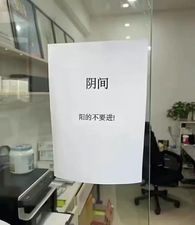 这是一张实拍的办公室场景图，画面主体是贴在办公区域玻璃门上的白色A4纸告示，告示上印有两行黑色文字，上方是“阴间”，下方是“阳的不要进!”。背景透过玻璃可以看到办公室内的打印机、办公座椅、置物架、绿植等常见办公设施。这张图运用了谐音梗，此处的“阴间”并非字面含义，而是指代新冠疫情期间专门留给抗原/核酸检测结果为阴性的员工使用的办公区域，以此幽默地提示检测结果为阳性的人员不要进入该空间，避免传染。