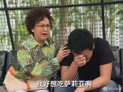 这是一张经过二次创作的TVB剧集《溏心风暴》截图梗图。画面中，穿着绿棕花纹衬衫、挎着米色挎包的中年女性（剧中角色荷妈）正伸手轻拍身旁年轻男性的肩膀，面露担忧地安抚他；年轻男性则捂脸低头，身体姿态显得十分崩溃委屈。图的下方添加了白色字幕“我好想吃萨莉亚啊”，将原本剧中安慰失意之人的伤感场景，改造成用夸张的崩溃情绪表达对平价西餐品牌萨莉亚的渴望，通过强烈的情绪反差制造出幽默搞笑的效果，精准戳中不少喜爱萨莉亚的网友的共鸣——萨莉亚以高性价比的西餐品类深受大众喜爱，用悲伤的失意场景放大想吃的心情，形成了极具网感的反差梗。