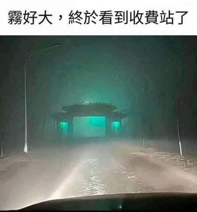 图片上方有文字“雾好大，終於看到收費站了”，下方是一张在浓雾中拍摄的道路照片。照片中，远处有一个被绿色灯光照亮的类似收费站的建筑结构，在浓重的雾气中显得有些神秘和模糊。道路两旁有路灯，但光线被雾气散射，能见度很低。整体画面营造出一种在恶劣天气中艰难行驶后终于看到目的地的感觉。
