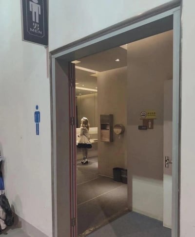 这是一张在男卫生间门口拍摄的照片，门外墙面有清晰的“男MEN”标识和蓝色男性图标，透过半开的门能看到，卫生间内的洗手区域站着一位金发的coser女孩，她穿着洛丽塔风格的女仆装类cos服装，背对着镜头。这种男厕出现女性装扮人物的场景形成了强烈的反差，容易引发观者的疑惑与好奇。
