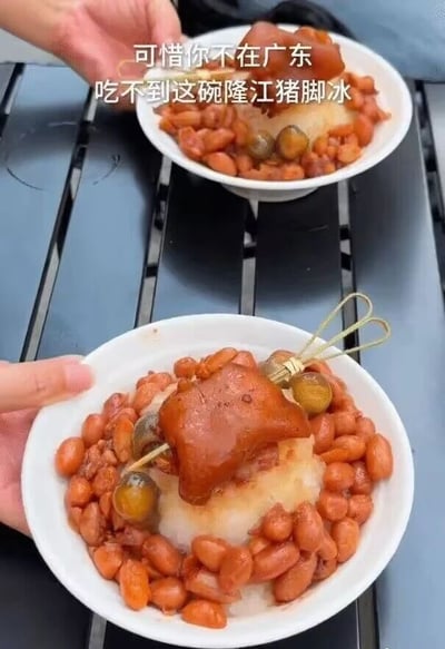 这是一张竖版美食实拍视频截图。画面中有两碗相同的食物，分别由两只手端着，放置在黑色条状桌面上。前景大碗最清晰，碗底是白色冰沙或牛奶冰，外围铺了一圈酱红色卤花生，中央放着一块体积较大、表皮酱红油亮的卤猪脚；猪脚旁还有串在竹签上的圆粒配料，视觉上类似蜜饯、水果或卤味配料。后景碗内容相同但略微虚化。画面上方叠加白色字幕：“可惜你不在广东 吃不到这碗隆江猪脚冰”。文案以地域安利的语气介绍这款广东网红猎奇食品，重点在于将隆江猪脚与冰品结合，形成强烈的甜咸、冷热反差。