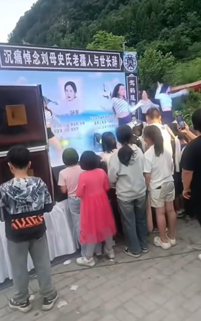 图片展示了一场户外葬礼现场，背景是山林。前方有一个大型悼念横幅，上面写着“沉痛悼念刘母史氏老人与世长辞”并配有逝者照片，显示享年58岁。横幅前有几位年轻女性正在进行舞蹈表演，台下有多名观众（包括儿童）在观看，部分人在用手机拍摄。这种在传统葬礼上进行娱乐表演的形式引发了关于丧葬文化和现代娱乐边界的讨论。