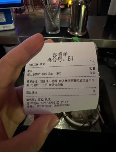 这张图是有人在酒吧吧台处手持展示一张顾客点单的客看单小票，小票显示桌台号为B1，就餐人数1人，原本点的菜品是一份售价30元的威士忌酸调酒，但整单备注内容十分荒诞：要求加蛋清不要酒，直接把酒换成红烧牛肉面，还要把蛋卧好，催促商家快点制作，小票的打印时间标注为2026年8月9日23点22分，背景可见吧台的金属容器、玻璃杯、收银设备等酒吧常见陈设，这份在酒吧点中餐的离谱订单形成了强烈的反差笑点。