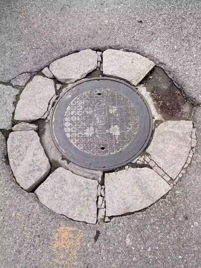 这是一张城市路面的实拍照片，画面中心是一块带有竖排“雨污”字样的圆形金属井盖，井盖周围的路面被切割成多块扇形石块，部分石块已经出现开裂、碎裂的情况，石块与井盖、外侧沥青路面之间存在明显缝隙，缝隙中还长出了少量杂草，直观呈现出市政路面设施老化、维护不足的状态。