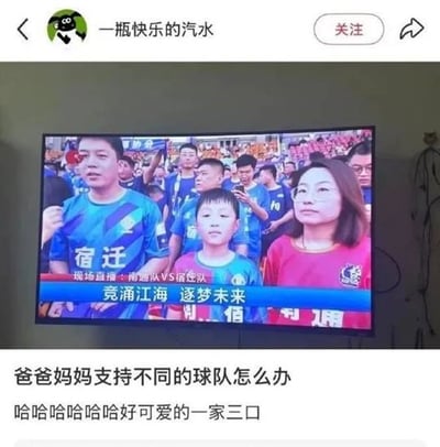 这是一张社交平台用户发布的内容截图，主体是用户实拍的电视播放的足球赛事直播画面，直播的是南通队对阵宿迁队的比赛，观众席里的一家三口着装十分有特点：父亲身穿蓝色宿迁队球迷服，母亲身穿红色南通队球迷服，中间的孩子则穿着红蓝拼接的球迷服，仿佛是父母支持不同球队的中和结果。发布者配文调侃提问爸爸妈妈支持不同的球队怎么办，还笑称这一家三口十分可爱。