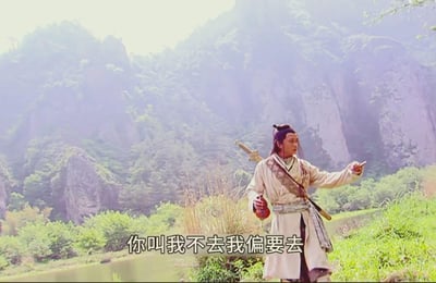 图中是2003版《倚天屠龙记》里的张无忌，他身着米白色古装，背负长剑站在青山绿水环绕的山间，手指前方，脸上带着不服气的神情，配文“你叫我不去我偏要去”，完美呈现出倔强叛逆、执意要做某事的状态，适合用来表达自己偏要违背他人劝阻的心态。