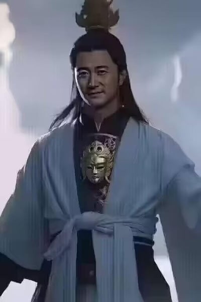 图片中是一位身着传统服饰的男子，他留着长发，头戴金色发冠，身穿蓝色长袍，胸前有金色面具状装饰。背景为朦胧的烟雾效果，整体呈现出一种古风仙侠或历史剧的氛围。男子面带微笑，姿态自然站立，眼神温和。
